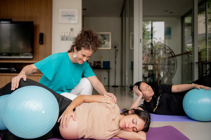 A pregnant woman exercising, lying on her left side, with Silvia helping her.