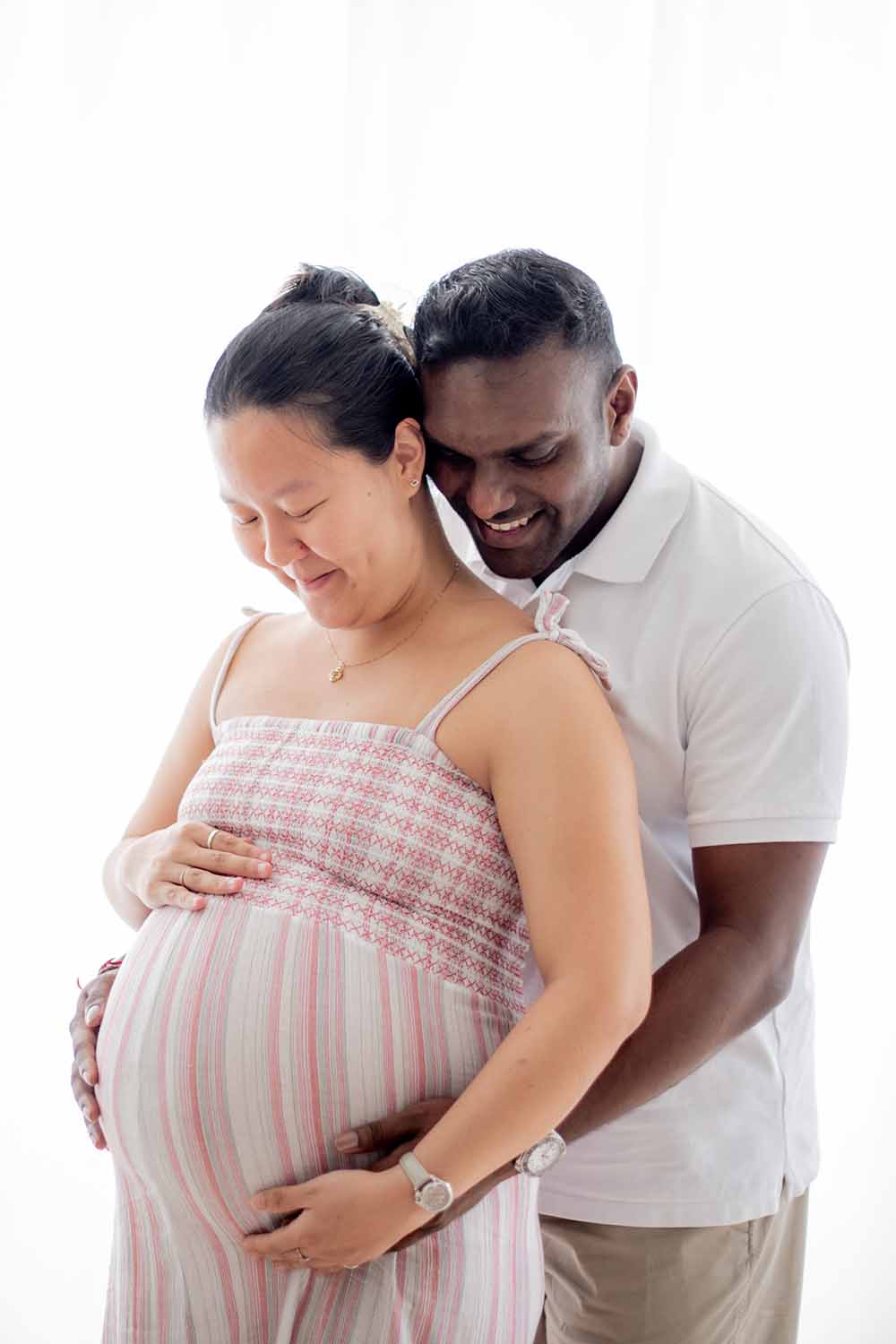 A pregnant woman, and her supportive husband standing behind her, and holding her.