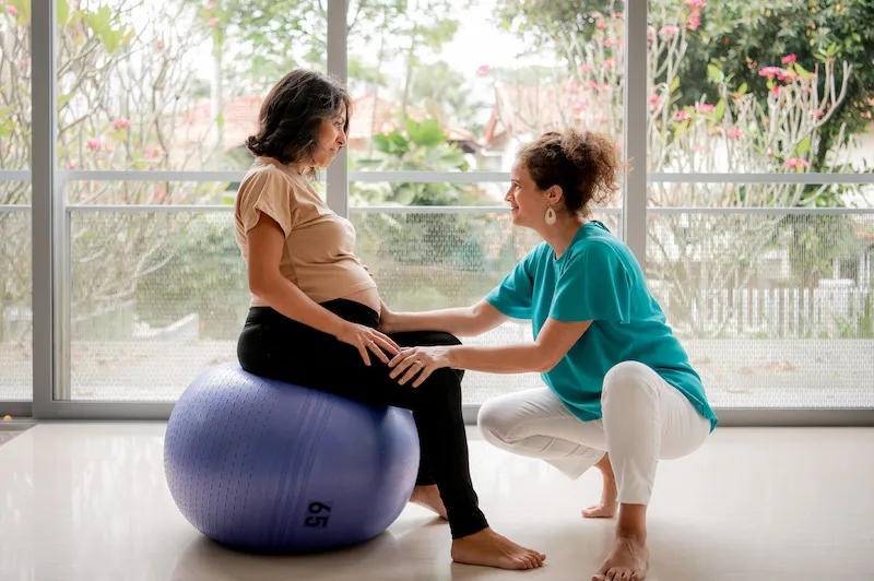 Silvia helping a pregnant woman exercise, as she sits on an exercise ball.